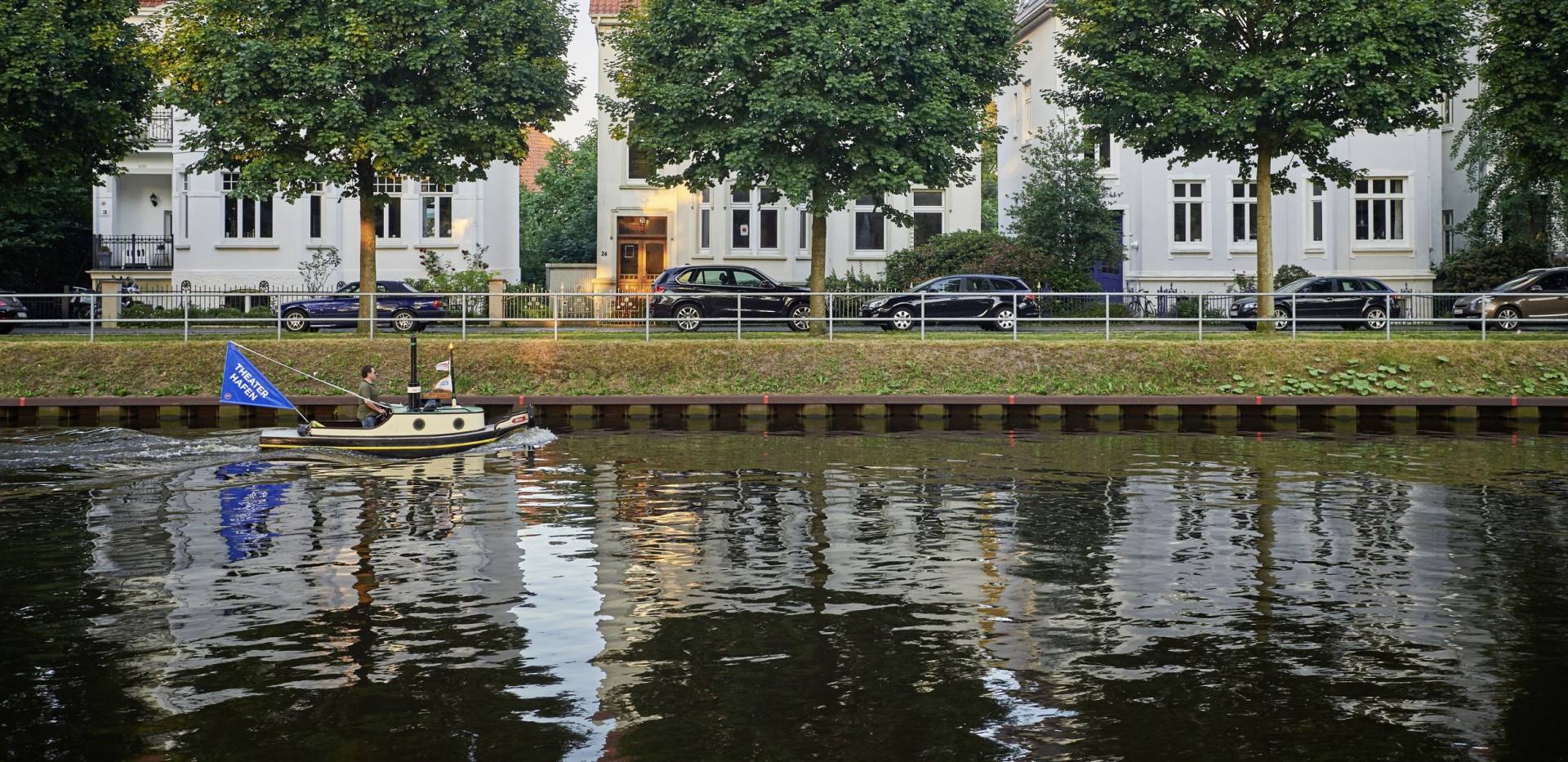 Ein kleines Boot f&auml;hrt auf dem K&uuml;stenkanal durch Oldenburg vor drei B&auml;umen und drei H&auml;usern.